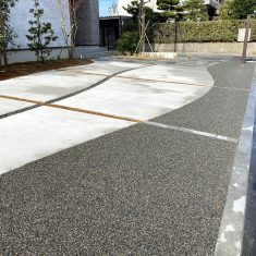 道路沿いの黒く見える箇所はバロンストーンという自然石の樹脂舗装材で、こちらにはお車の乗り入れが可能なタイプの商品を使用しています。駐車場には、芝・インターロッキング・大磯砂利の3種類の目地を設けています。奥の方に見える道路境界付近にはブロックとフェンスを施工しています。